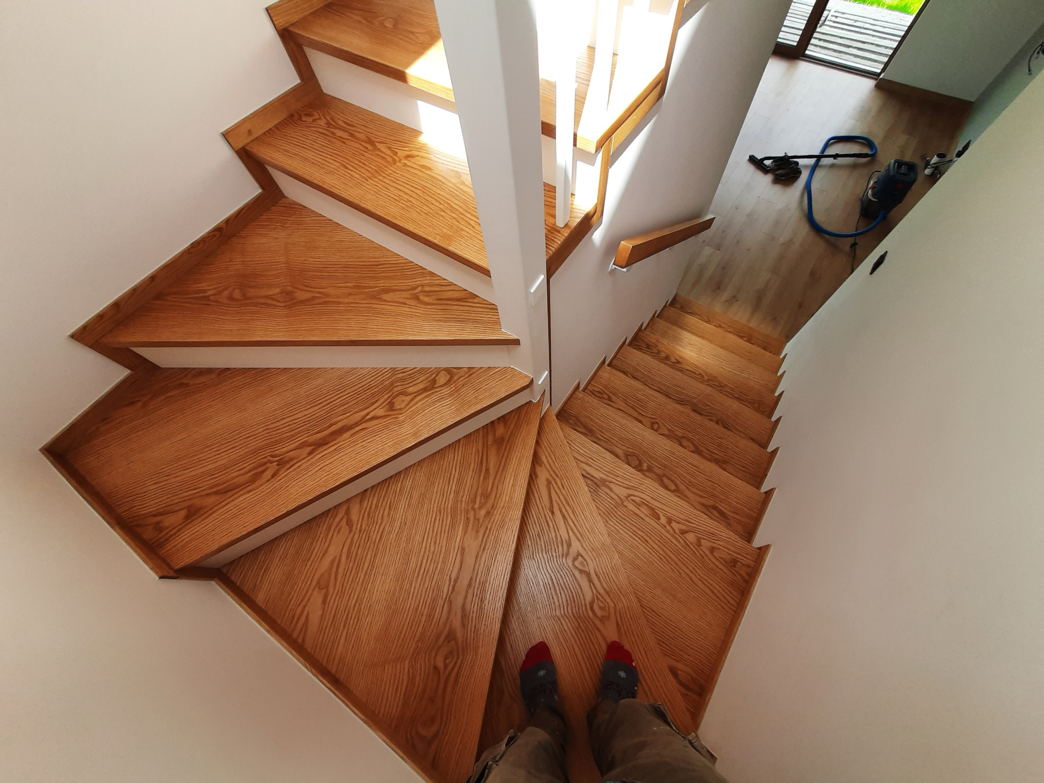 Drewniane schody z białymi barierkami, widok z góry na stopnie o trójkątnym kształcie. Na dole kadru widoczne stopy w skarpetach. W tle odkurzacz.