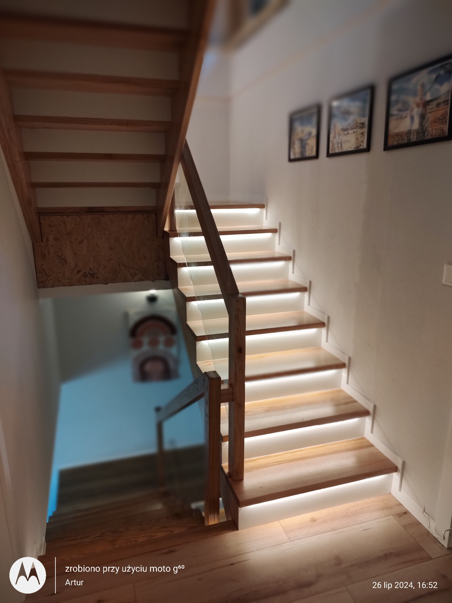 Drewniane schody z podświetleniem LED, widok z dołu. Balustrada z drewna i szkła. Na ścianie wiszą obrazy w czarnych ramkach. Widoczne drewniane wykończenie pod schodami.