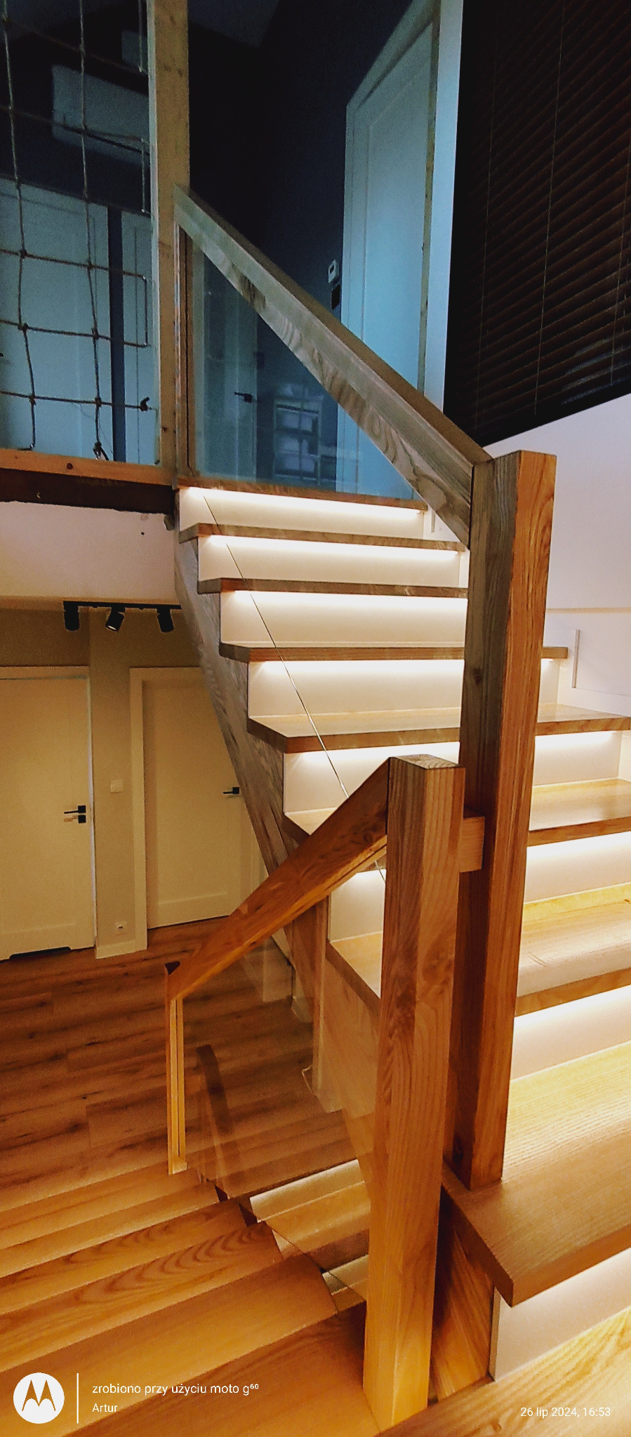 Drewniane schody z podświetleniem LED i szklaną balustradą. Widoczne detale wykończenia i naturalna kolorystyka drewna. Ujęcie z dołu, podkreślające konstrukcję.