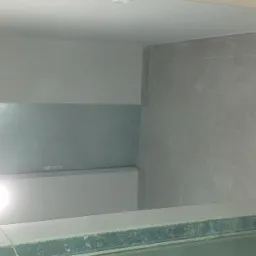 Wnętrze łazienki w trakcie remontu, widoczne szare płytki na podłodze i ścianach, fragment ściany przygotowanej do dalszych prac, tymczasowe oświetlenie halogenowe.