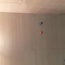 Ściana łazienki wyłożona beżowymi płytkami ceramicznymi z widocznymi otworami na instalację wodną i częściowo ułożoną ciemną podłogą, ukazująca postęp prac glazurniczych.