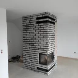 Murowany kominek z szarych cegieł z czarną fugą w surowym wnętrzu, widoczne kable i niedokończone ściany.