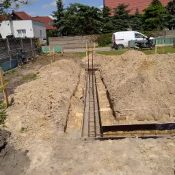 Przygotowanie fundamentów pod budowę domu: wykopany rów z szalunkiem i zbrojeniem stalowym, ułożony w ziemi, na tle placu budowy z zaparkowanym białym busem i skuterem.