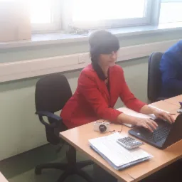 Kobieta w czerwonej marynarce pracuje przy biurku na laptopie, obok leży kalkulator, dokumenty i aparat fotograficzny.