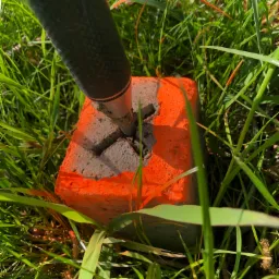 Pomarańczowy słupek geodezyjny z wyżłobionym krzyżem, wbity w ziemię porośniętą trawą, z widocznym fragmentem tyczki geodezyjnej.