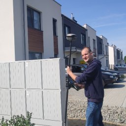 Usługi Geodezyjne GEO-JAW Michał Pacut - Mężczyzna w bluzie i jeansach, uśmiechnięty, trzyma tyczkę geodezyjną z odbiornikiem GPS na tle szeregu nowoczesnych domów jednorodzinnych i skrzynki pocztowej.