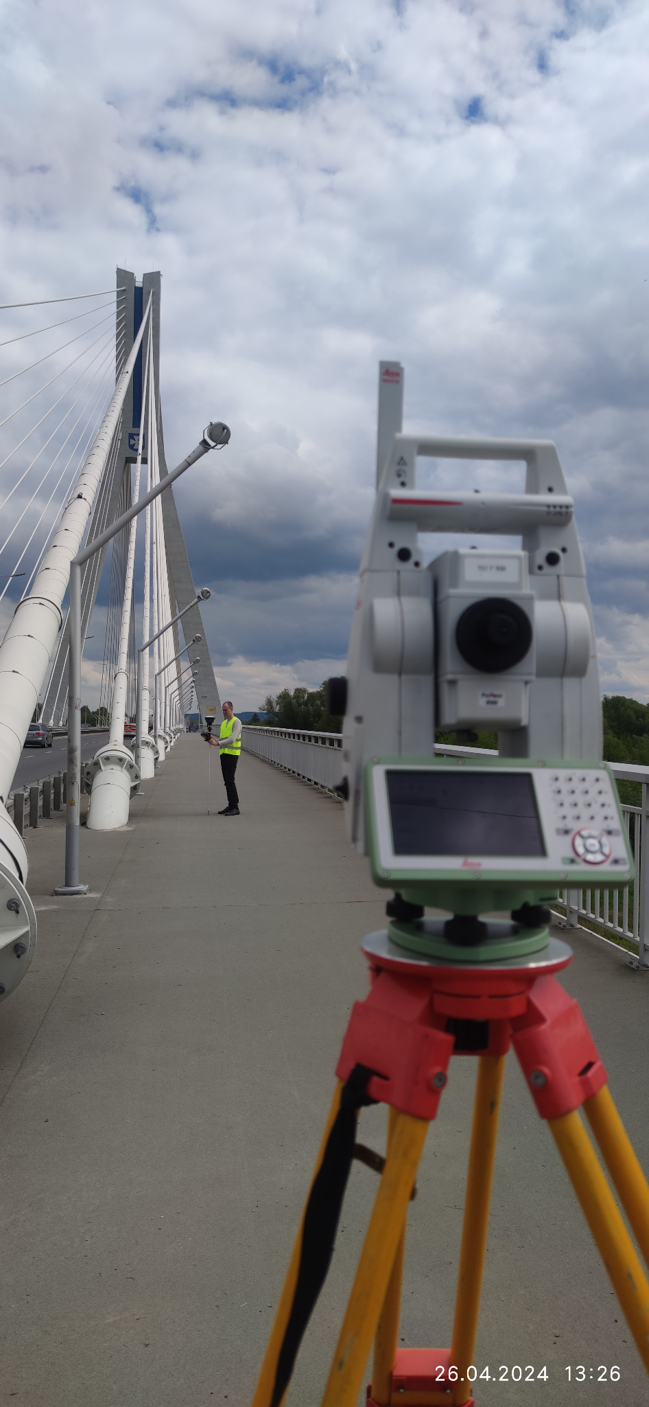 Pomiar mostu linowego za pomocą tachimetru Leica, pracownik w żółtej kamizelce odblaskowej z tyczką geodezyjną, pochmurne niebo w tle.