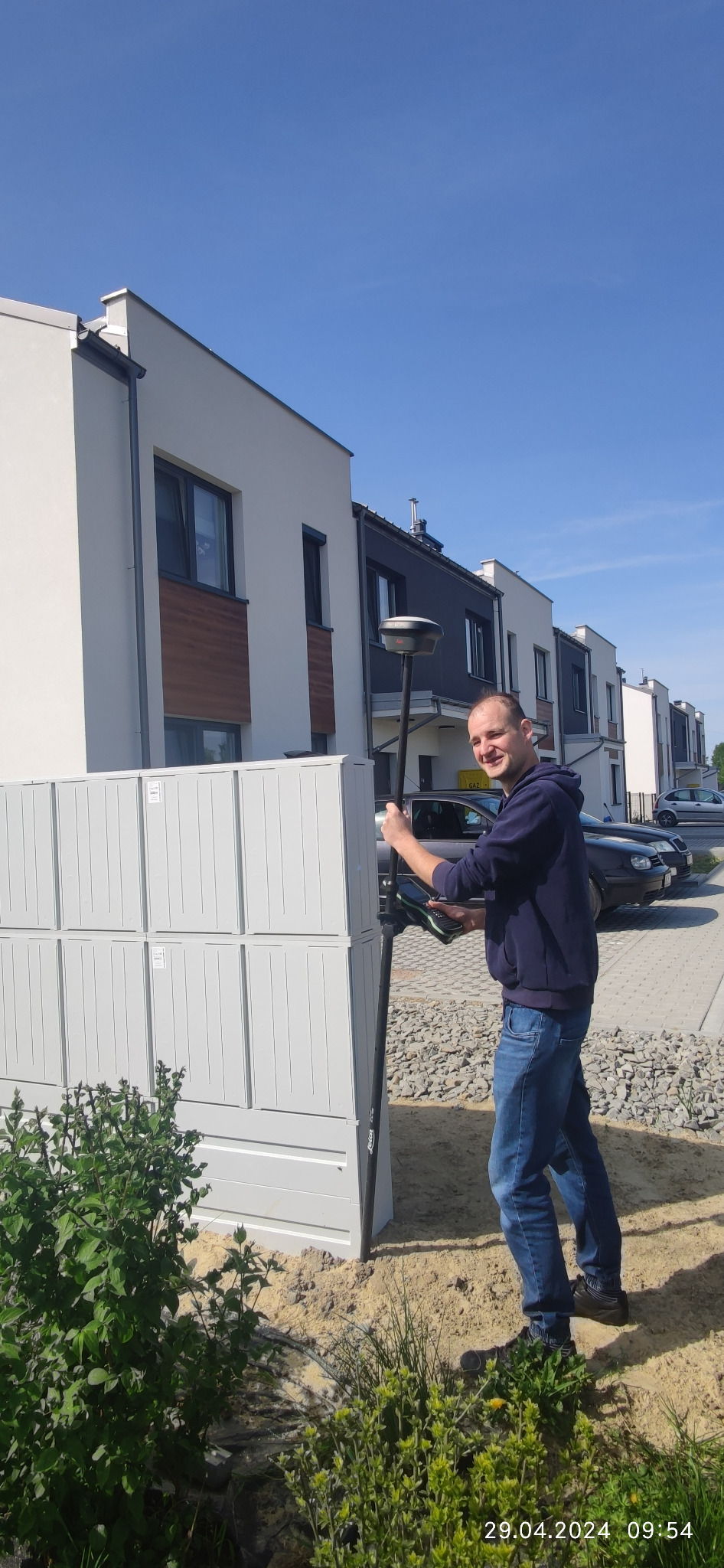 Mężczyzna w bluzie i jeansach, uśmiechnięty, trzyma tyczkę geodezyjną z odbiornikiem GPS na tle szeregu nowoczesnych domów jednorodzinnych i skrzynki pocztowej.