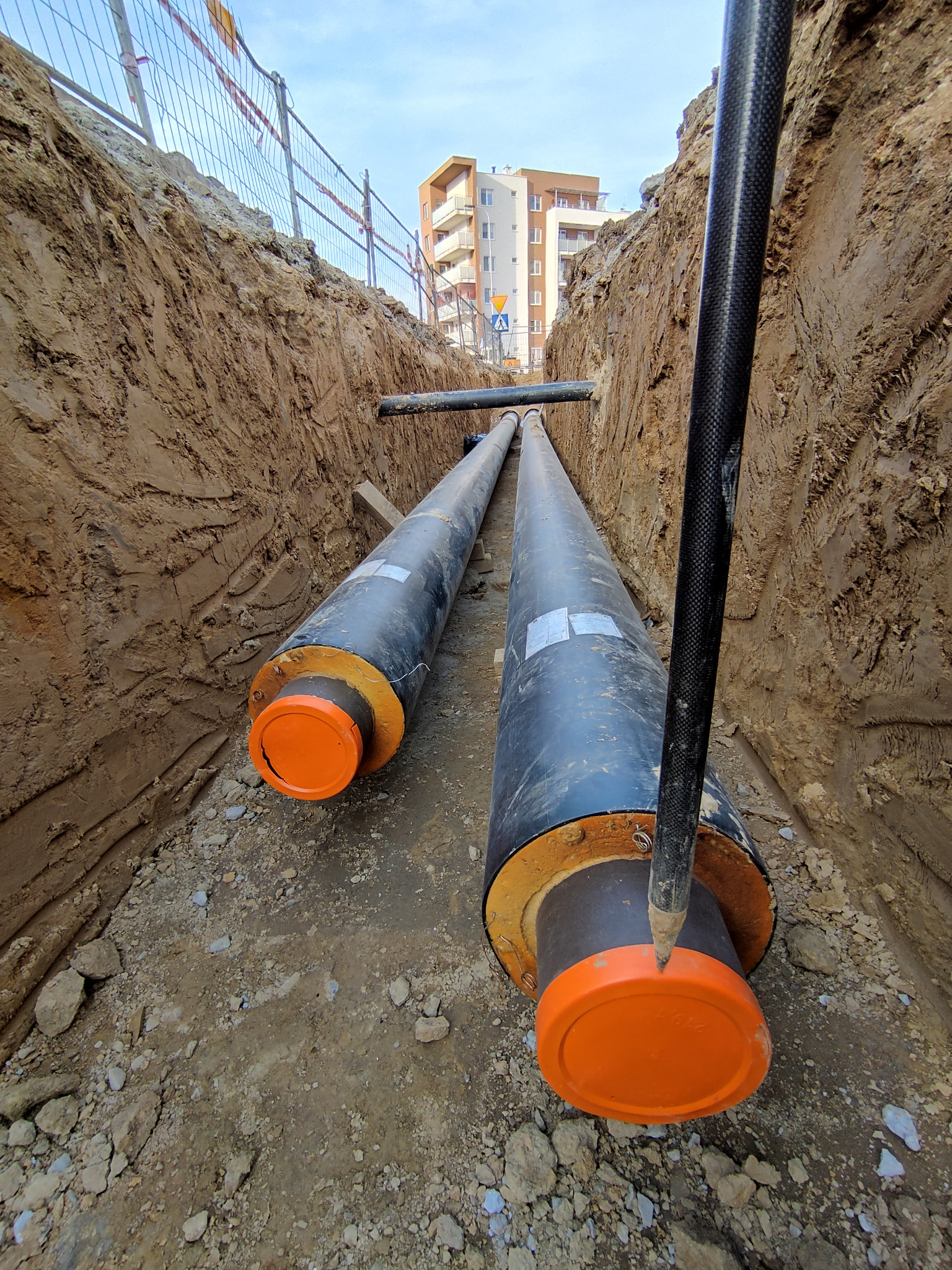Dwa rurociągi ciepłownicze z pomarańczowymi zaślepkami ułożone w głębokim wykopie ziemnym na tle budynku mieszkalnego i metalowego ogrodzenia.
