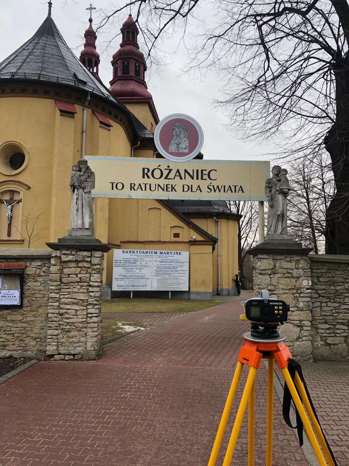 Pomarańczowy niwelator na trójnogu ustawiony na brukowanym chodniku przed żółtym budynkiem kościoła z czerwonymi wieżami i tablicą 'Różaniec to ratunek dla świata'.