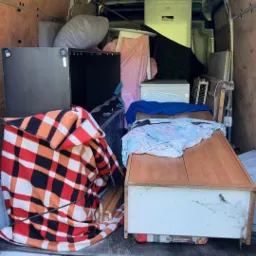 Wnętrze częściowo załadowanego busa z meblami, sprzętem AGD i kocami w kratę, widok z zewnątrz przez otwarte tylne drzwi.