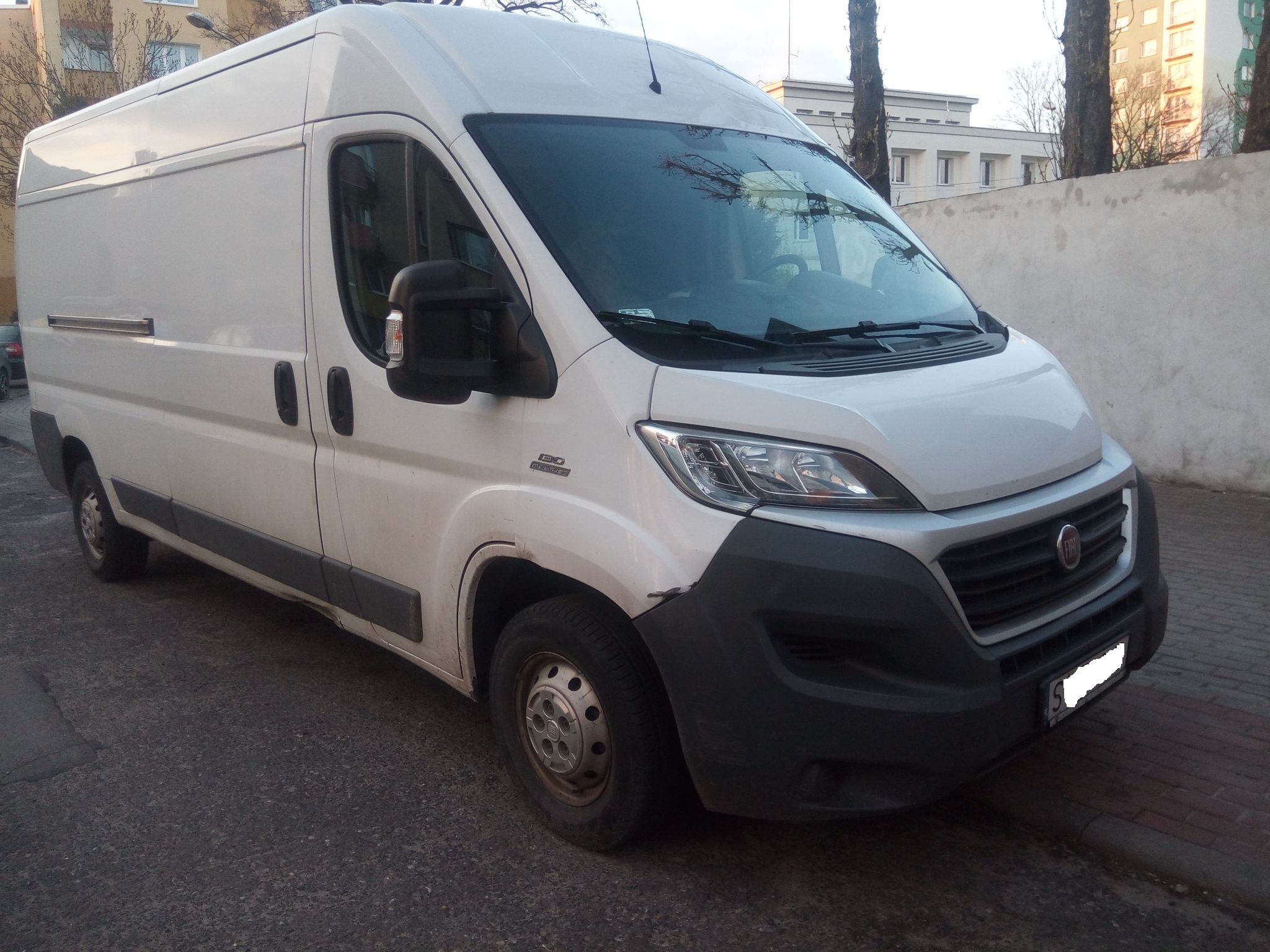 Biały Fiat Ducato, widok z przodu, zaparkowany na ulicy w pobliżu chodnika i ściany, widoczne ślady użytkowania i niewielkie zabrudzenia.