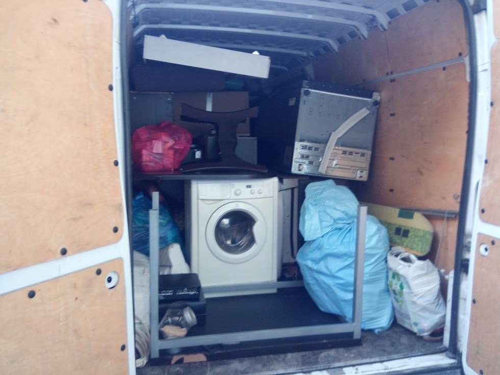 Wnętrze białego busa załadowane różnymi przedmiotami, w tym pralką, meblami i zapakowanymi torbami, zabezpieczonymi metalową ramą, widoczne drewniane ściany pojazdu.