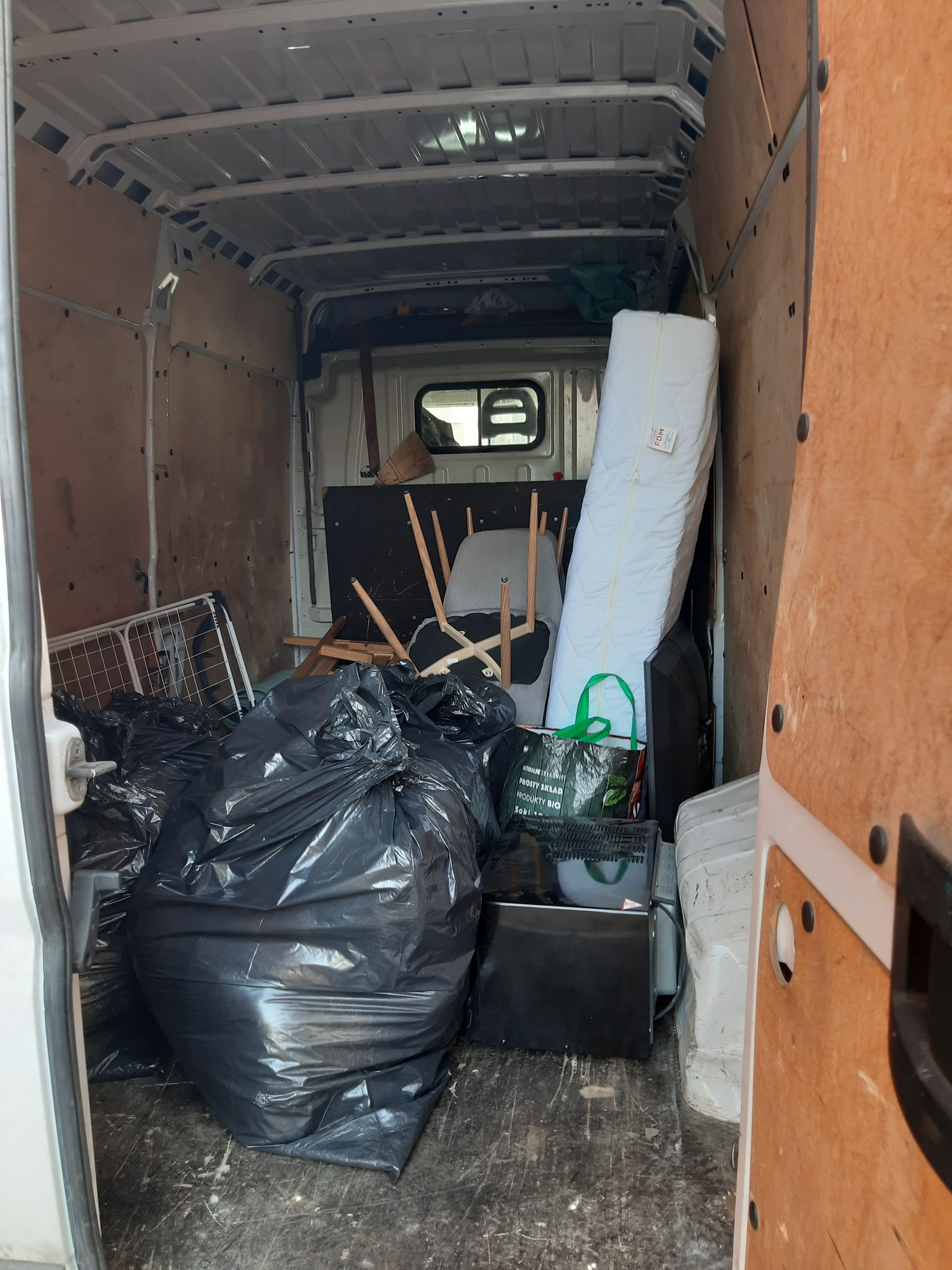 Wnętrze furgonetki załadowane workami ze śmieciami, rozłożonymi krzesłami, materacem i sprzętem RTV podczas przeprowadzki.