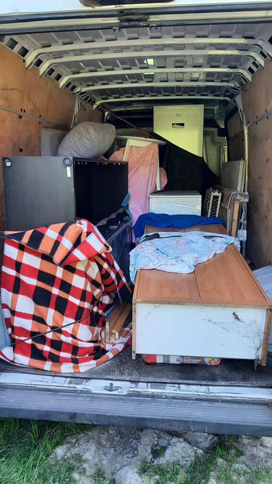 Wnętrze częściowo załadowanego busa z meblami, sprzętem AGD i kocami w kratę, widok z zewnątrz przez otwarte tylne drzwi.