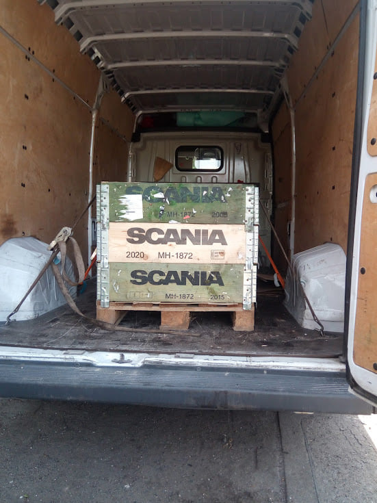 Wnętrze furgonetki z załadowanymi, drewnianymi skrzyniami na palecie, zabezpieczonymi pasami transportowymi. Skrzynie oznaczone logiem SCANIA i datami produkcji.