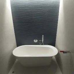 Nowoczesna łazienka w trakcie wykończenia: wolnostojąca biała wanna owalna na tle ściany z ciemnych, poziomych płytek imitujących fale, widoczne przyłącza wodne z zaślepkami.