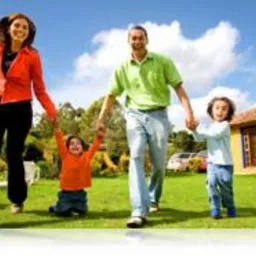 Uśmiechnięta rodzina z dwójką dzieci spaceruje po zielonym trawniku na tle błękitnego nieba z lekkimi chmurami; w tle widoczny budynek i zaparkowany samochód.