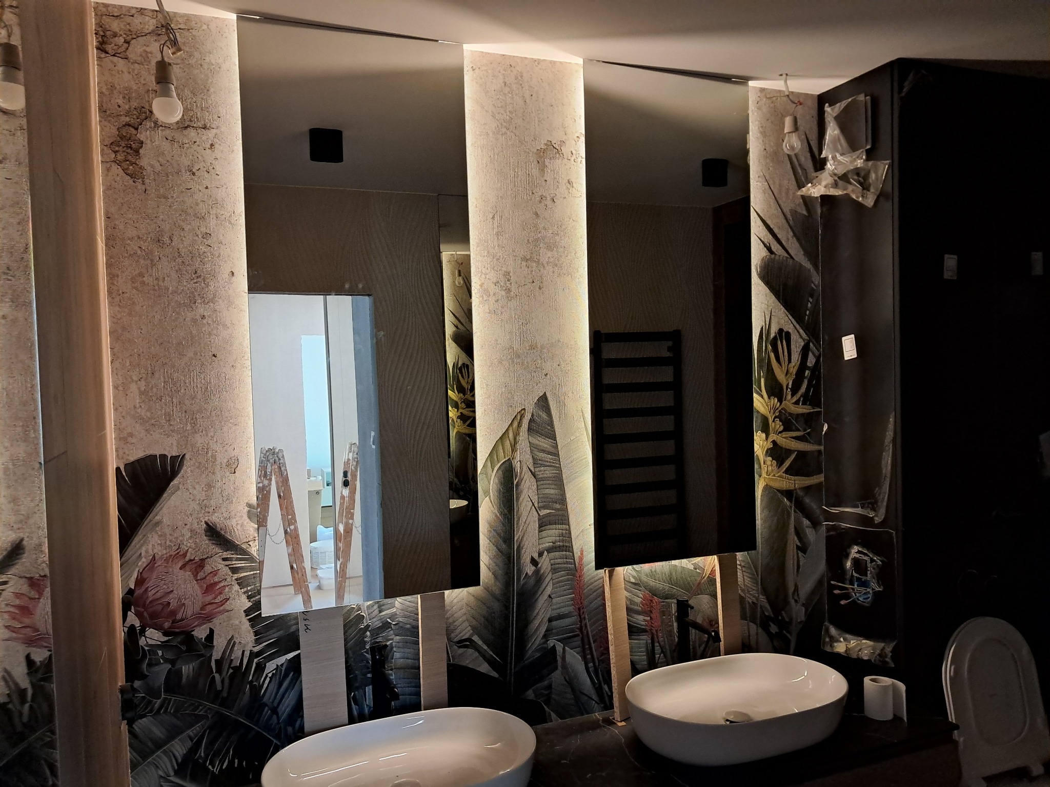 Nowoczesna łazienka po remoncie z dwoma umywalkami, lustrami z podświetleniem i motywem roślinnym na ścianach. Widoczne drabiny malarskie.