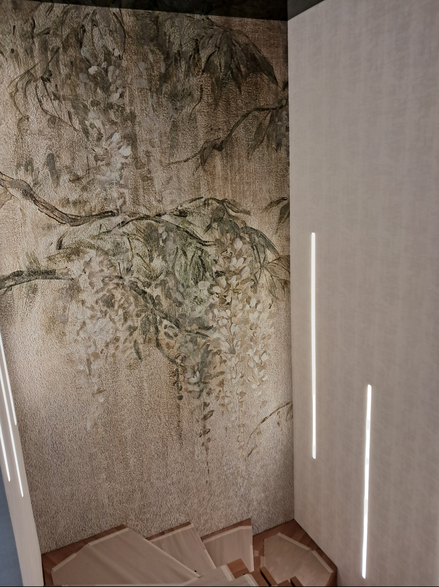 Ściana z dekoracyjną tapetą w odcieniach beżu i brązu z motywem roślinnym, oświetlona pionowymi pasami światła, widoczne fragmenty schodów.