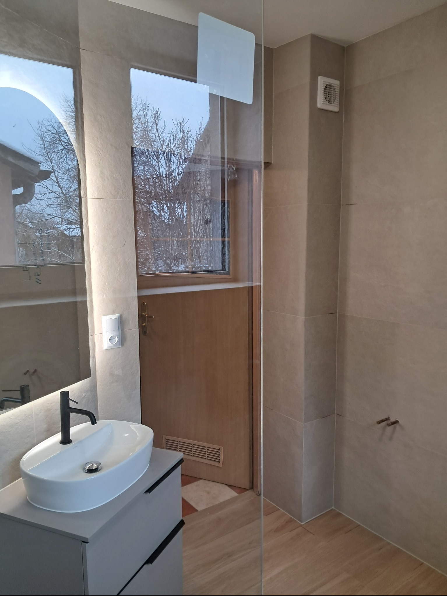 Nowoczesna łazienka z owalną umywalką na szafce, przeszklonym prysznicem i beżowymi płytkami. Okno z widokiem na zimowy krajobraz dodaje naturalnego światła.