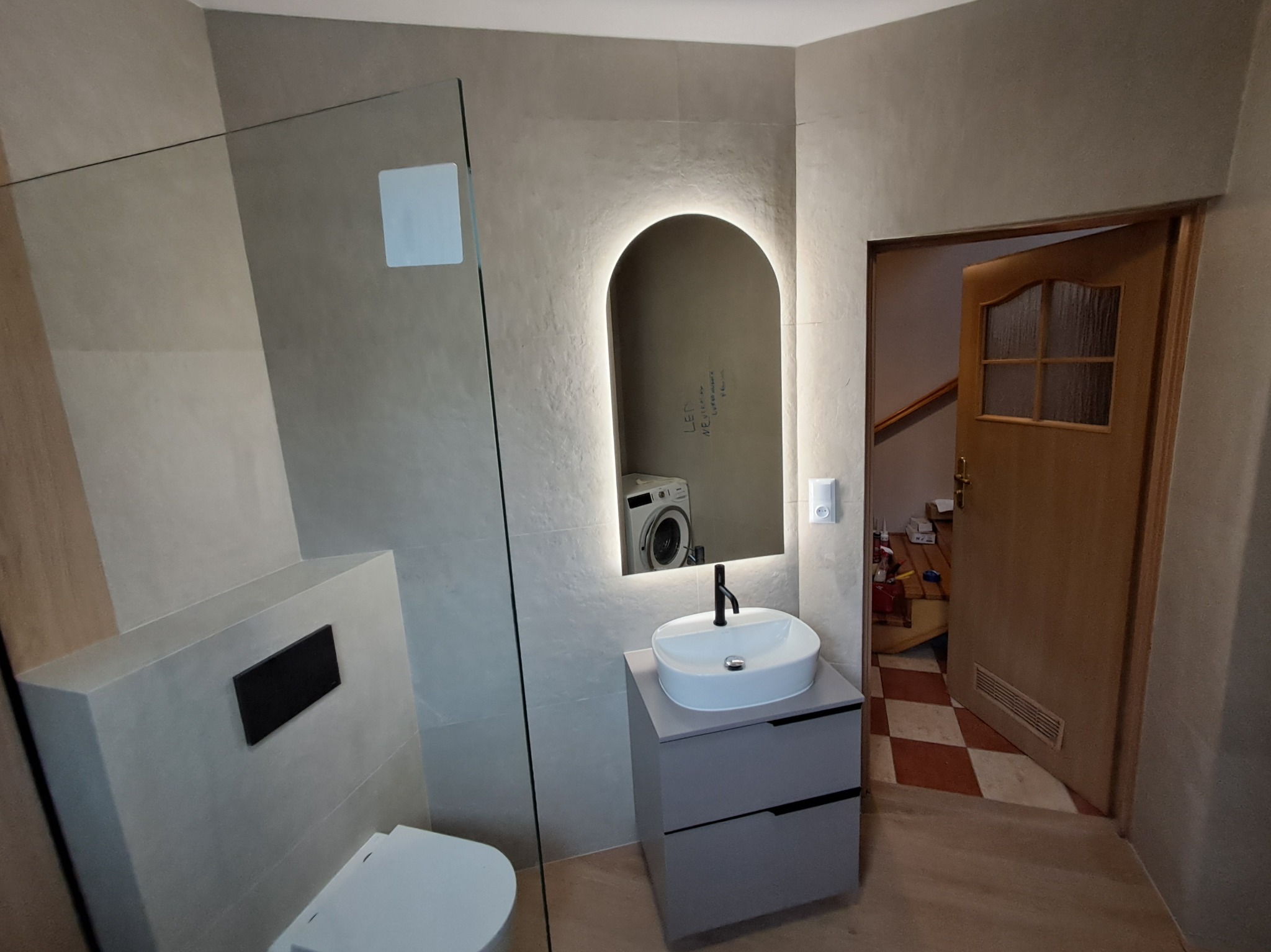 Nowoczesna łazienka z kabiną prysznicową, umywalką na szafce i podświetlanym lustrem. Uwagę przyciąga pralka widoczna w odbiciu lustra.