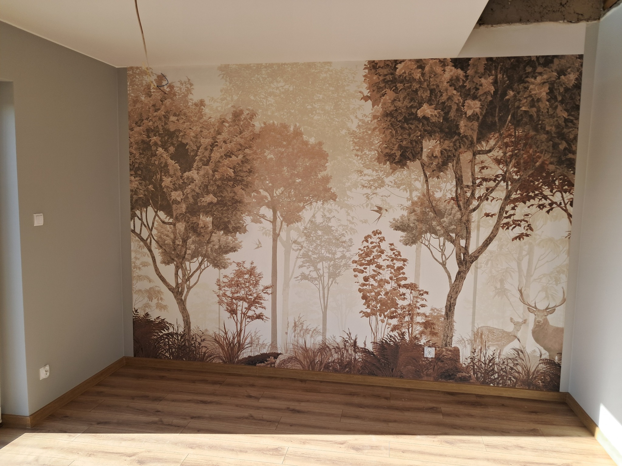 Pokój z fototapetą leśną w odcieniach brązu. Na podłodze panele, ściany szare. Fragment sufitu z widocznymi niedoróbkami. Realizacja z dbałością o detal.