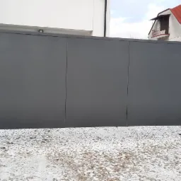 Nowoczesna, przesuwna brama wjazdowa w kolorze grafitowym, z widocznymi betonowymi słupkami i fragmentami instalacji elektrycznej, na tle zaśnieżonego podjazdu i budynku z czerwoną dachówką.