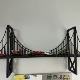 Metalowa półka w kształcie mostu wiszącego z zabawkami samochodami na tle białej ściany, widoczne magnesy na lodówce w dolnej części kadru.