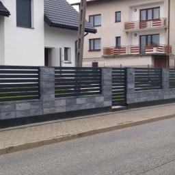 STAR-SPAW Ziejka Mariusz - Nowoczesne ogrodzenie z szarej kostki i poziomych, ciemnych przęseł, z furtką pośrodku, na tle zabudowy jednorodzinnej.