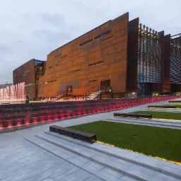 Nowoczesny budynek z rdzawą elewacją, fontanną z czerwonym podświetleniem i zielonymi trawnikami, widok z dołu na tle pochmurnego nieba.