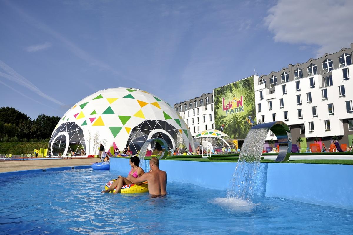 Para relaksuje się w basenie na dmuchanym kole w parku wodnym z kopułami geodezyjnymi i wodospadami ze stali nierdzewnej, na tle budynku z napisem Lemon Park.