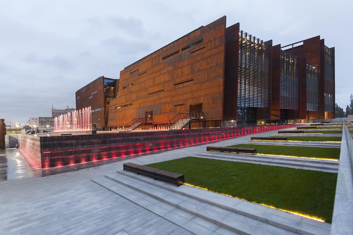 Nowoczesny budynek z rdzawą elewacją, fontanną z czerwonym podświetleniem i zielonymi trawnikami, widok z dołu na tle pochmurnego nieba.