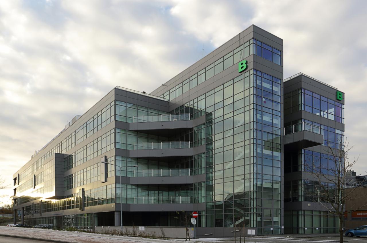 Nowoczesny budynek biurowy z przeszkloną fasadą i balkonami, oświetlony popołudniowym słońcem, z widocznym logo 'B' na elewacji.