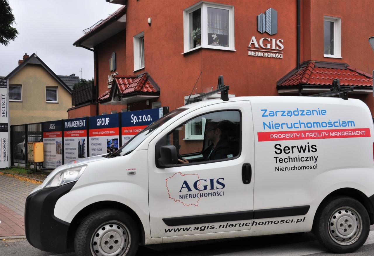 Biały samochód dostawczy z logo i hasłami firmy zajmującej się zarządzaniem nieruchomościami na tle budynku biurowego z logo tej samej firmy.