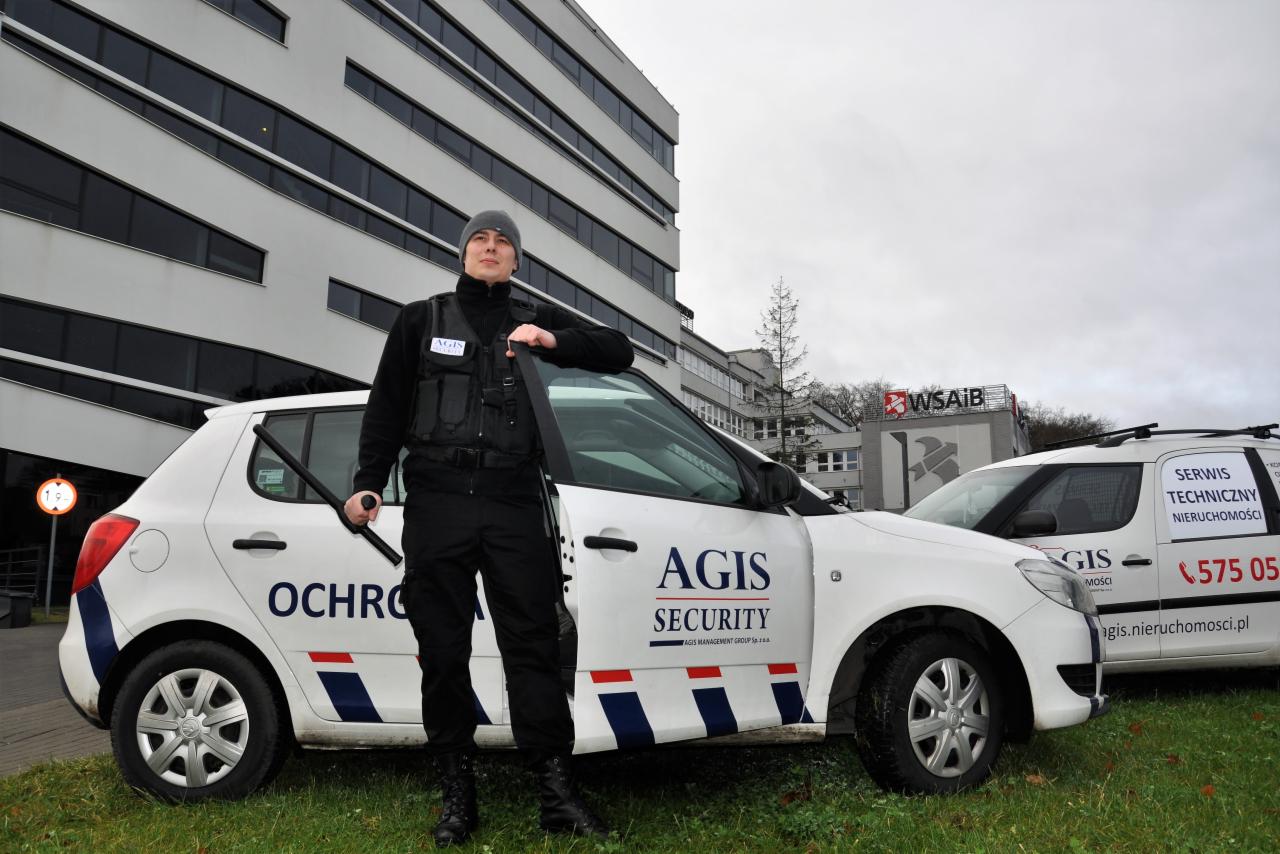 Ochroniarz w czarnym uniformie i czapce, oparty o biały samochód z napisem AGIS SECURITY, w tle budynek biurowy z dużą ilością okien.