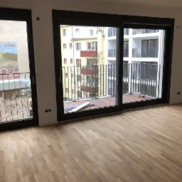 Pusty pokój z parkietem i dużymi oknami balkonowymi z czarnymi ramami, widok na balkony sąsiednich budynków.