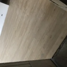 Ułożona podłoga z paneli drewnopodobnych w jasnym odcieniu, widoczna krawędź z surowym betonem i fragment białej ściany z dwoma otworami na gniazdka elektryczne.