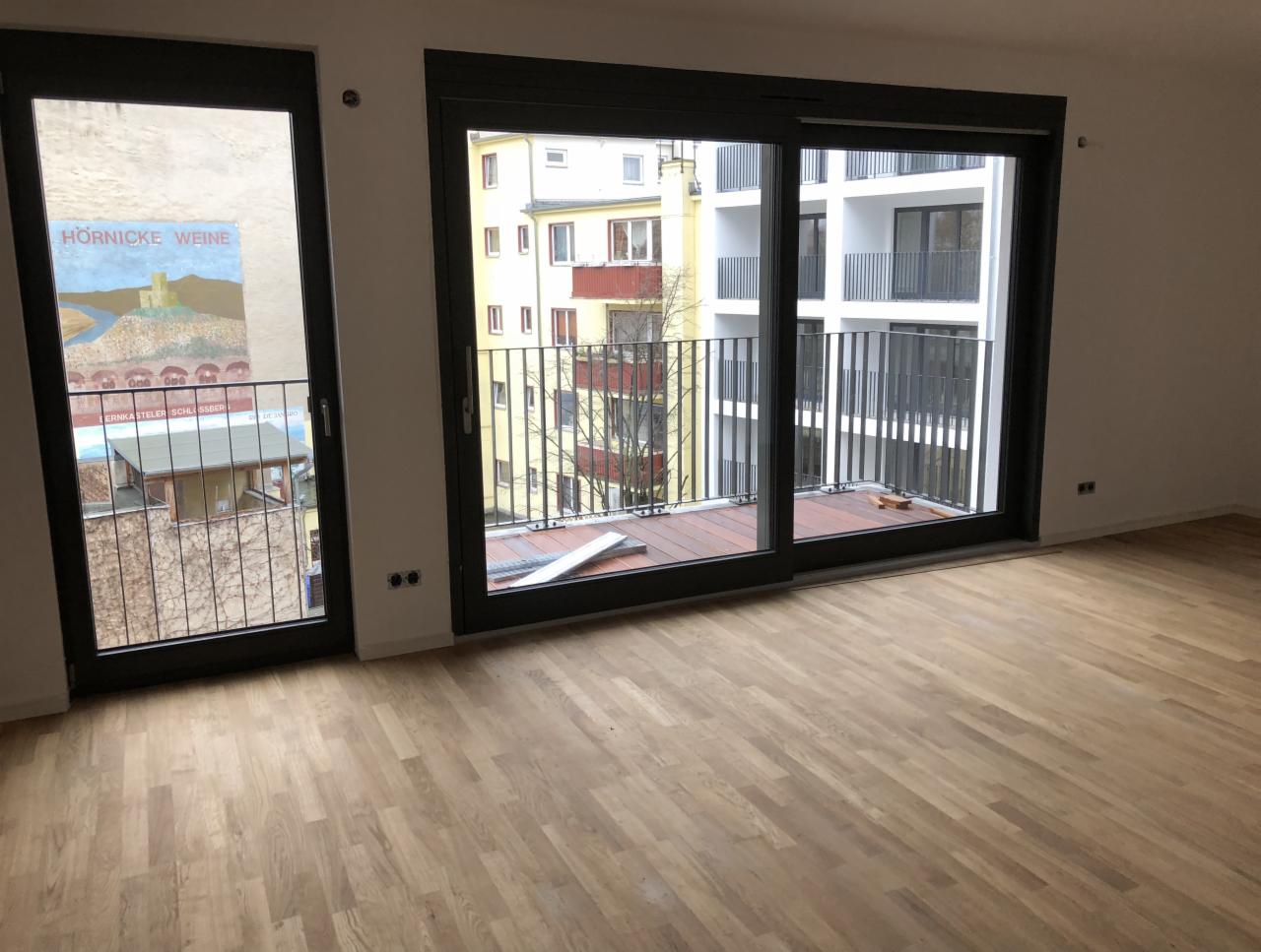 Pusty pokój z parkietem i dużymi oknami balkonowymi z czarnymi ramami, widok na balkony sąsiednich budynków.