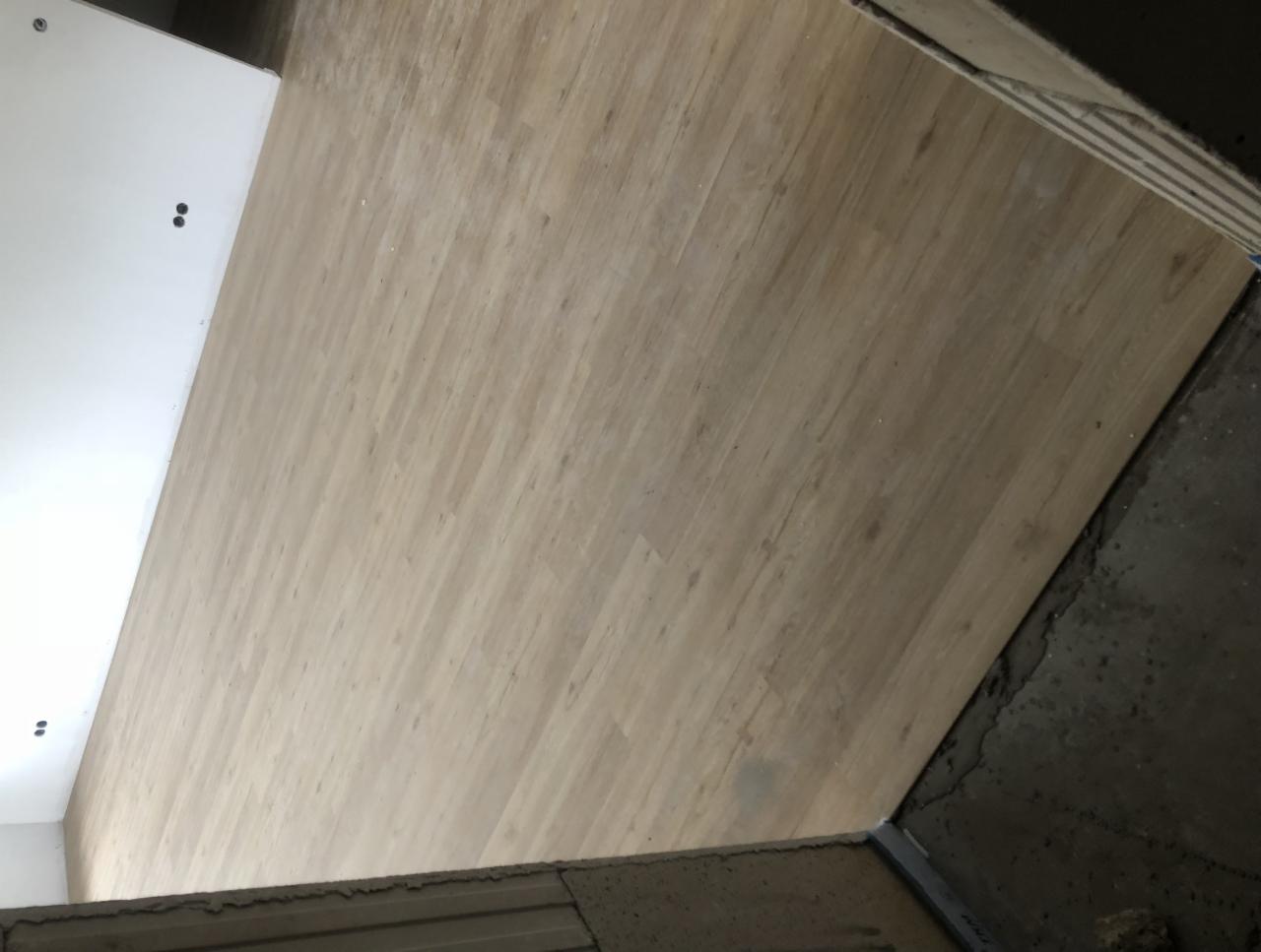 Ułożona podłoga z paneli drewnopodobnych w jasnym odcieniu, widoczna krawędź z surowym betonem i fragment białej ściany z dwoma otworami na gniazdka elektryczne.