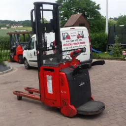 Czerwony wózek paletowy elektryczny Linde L12 na kostce brukowej, z widocznym samochodem dostawczym z reklamą w tle i elementami ogrodu. Ujęcie z bliska, lekko z dołu.