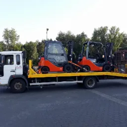 Dwa pomarańczowe wózki widłowe Toyota zabezpieczone pasami transportowymi na żółtej lawecie białej ciężarówki z widocznym logo firmy na drzwiach.