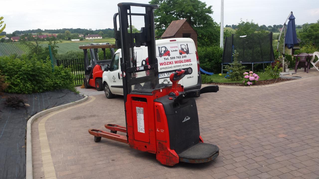 Czerwony wózek paletowy elektryczny Linde L12 na kostce brukowej, z widocznym samochodem dostawczym z reklamą w tle i elementami ogrodu. Ujęcie z bliska, lekko z dołu.