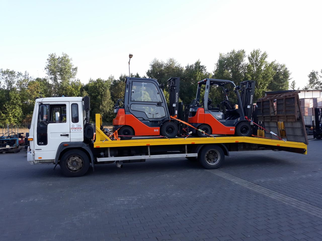 Dwa pomarańczowe wózki widłowe Toyota zabezpieczone pasami transportowymi na żółtej lawecie białej ciężarówki z widocznym logo firmy na drzwiach.