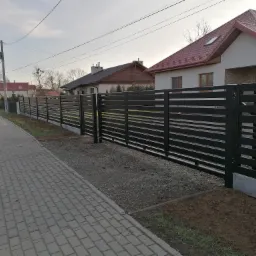 Nowoczesne, poziome, czarne ogrodzenie z metalowych profili na tle domów jednorodzinnych i chodnika z kostki.