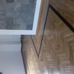 Parkiet ułożony w jodełkę z czarnymi paskami dekoracyjnymi, widoczna część ściany i fragment kamiennej okładziny.