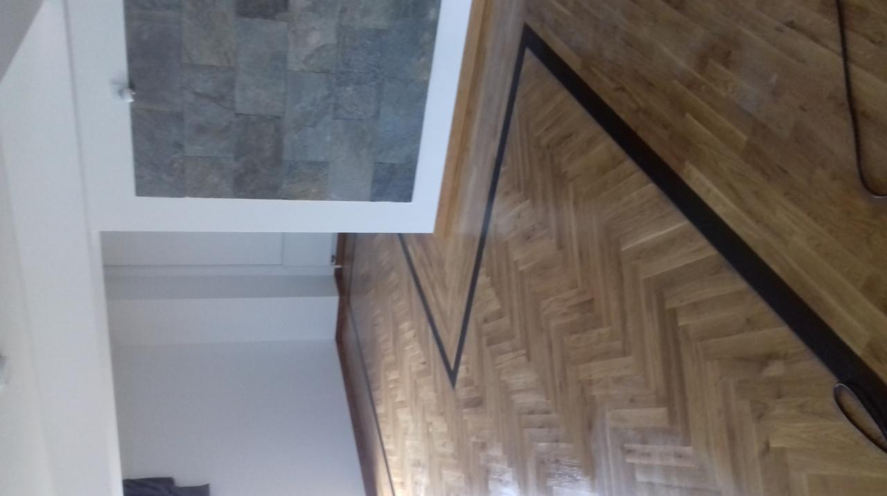 Parkiet ułożony w jodełkę z czarnymi paskami dekoracyjnymi, widoczna część ściany i fragment kamiennej okładziny.