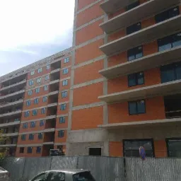 Budynek wielorodzinny w trakcie budowy, widoczne zamontowane ramy okienne, surowy stan elewacji z cegły i betonu, ogrodzenie budowy w pierwszym planie.