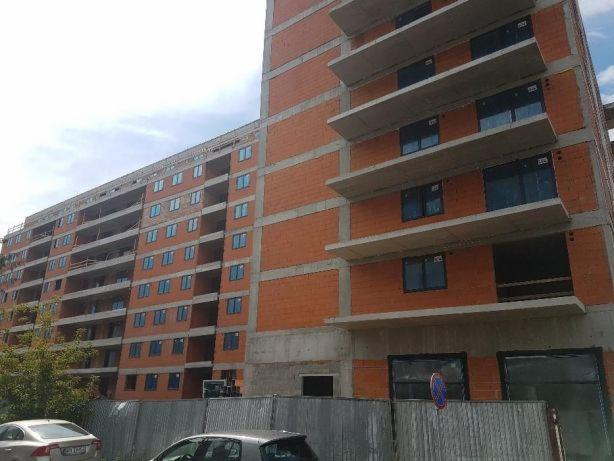 Budynek wielorodzinny w trakcie budowy, widoczne zamontowane ramy okienne, surowy stan elewacji z cegły i betonu, ogrodzenie budowy w pierwszym planie.