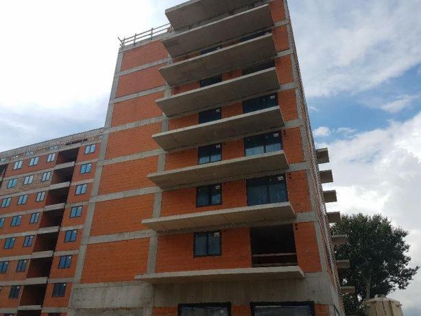 Budynek w trakcie budowy z widocznymi otworami okiennymi i betonowymi balkonami, ceglana elewacja w surowym stanie, widoczne niebo i drzewa w tle.